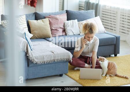 Jeune femme avec des cheveux bouclés assis sur le sol avec son chat et utilisant un ordinateur portable dans la chambre à la maison Banque D'Images