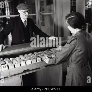 1950s, homme âgé dans une chemise, cravate et casquette plate, parlant à une bibliothécaire se tenant devant des plateaux en bois de fiches dans cette image historique de J Allan Cash. Banque D'Images