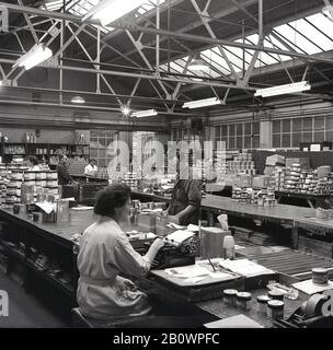 Années 1950, historique, dans une usine de peinture, les travailleurs masculins et féminins dans un haut plafond rempli de lumière d'entrepôt d'emballage et de traitement des commandes pour la livraison, avec une travailleuse assise à un long établi utilisant une machine à écrire, Angleterre, Royaume-Uni. Banque D'Images