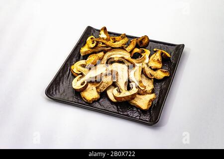 Champignons porcini séchés isolés sur fond blanc. Ingrédient traditionnel pour cuisiner des aliments sains. Plaque en céramique noire, espace de copie Banque D'Images