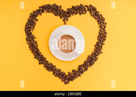 Tasse de café blanche, grains de café en forme de coeur, fond jaune. Vue de dessus. Pose plate. Espace de copie. Banque D'Images