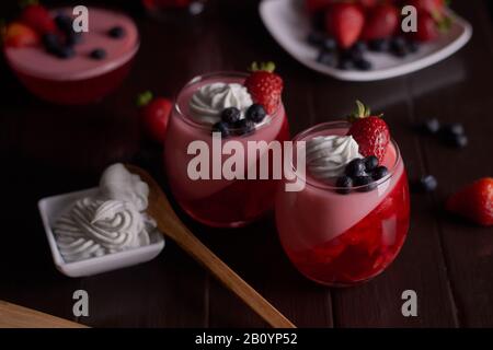 Desserts à base de gelée de fruits rouges avec crème chantilly dans des tasses en verre avec bleuets et tasse en céramique blanche avec crème chantilly avec fond de bois de café Banque D'Images