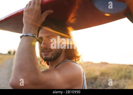 Photo d'un homme dérasé et content regardant un appareil photo tenant le surf sur sa tête à la plage ensoleillée Banque D'Images