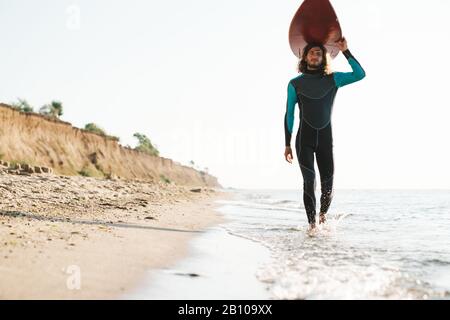 Photo de l'homme masculin athlétique en combinaison tenant planche de surf sur sa tête tout en marchant sur la plage ensoleillée Banque D'Images