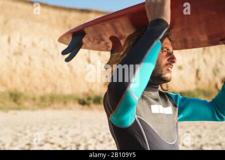 Photo de l'homme barbu athlétique en combinaison tenant planche de surf sur sa tête tout en marchant sur la plage ensoleillée Banque D'Images