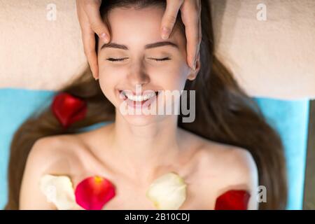 Jeune belle femme recevant un massage du visage de tête dans un spa de beauté avec pétales de fleur de rose Banque D'Images