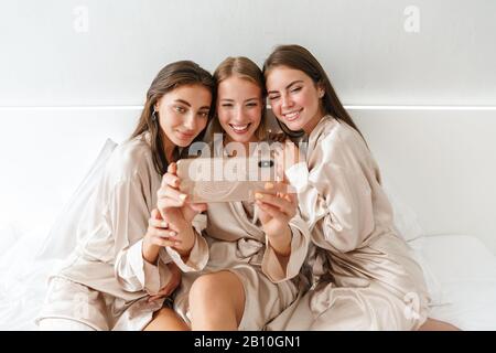 Image de jeunes filles heureuses femmes amis à l'intérieur sur le lit à la fête de poule à la maison prendre un selfie par téléphone portable. Banque D'Images