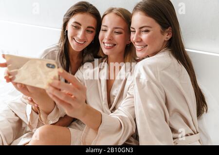 Image de jeunes filles heureuses femmes amis à l'intérieur sur le lit à la fête de poule à la maison prendre un selfie par téléphone portable. Banque D'Images