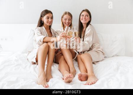 Trois jeunes filles amies portant des robes de chambre assis sur le lit à la maison, boire du champagne, faire la fête Banque D'Images