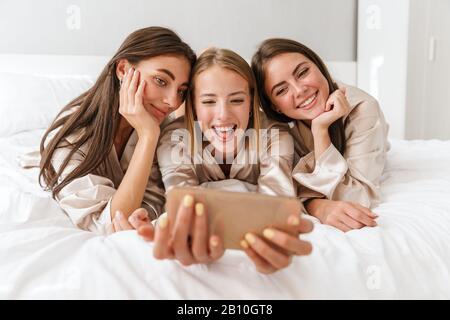 Trois filles gaies amis portant des robes de chambre pontant sur le lit à la maison, prenant un selfie Banque D'Images