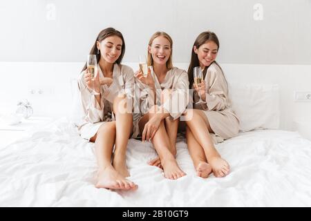 Trois jeunes filles amies portant des robes de chambre assis sur le lit à la maison, boire du champagne, faire la fête Banque D'Images