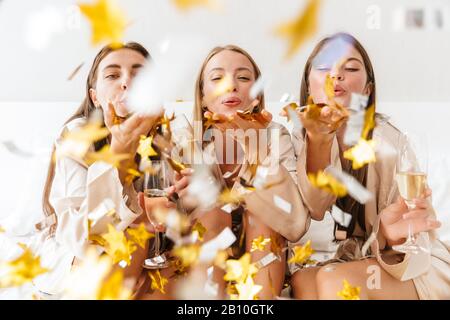 Trois jeunes filles amies portant des robes de chambre assis sur le lit à la maison, boire du champagne, faire la fête, souffler des confettis Banque D'Images