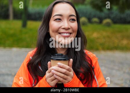 Jeune femme asiatique souriante portant un imperméable marchant à l'extérieur sous la pluie, tenant une tasse de café, regardant loin Banque D'Images