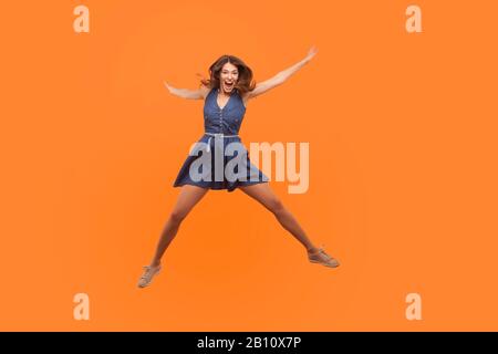 Une femme de brunette exstatic surplaisante et excitée, vêtue d'une robe en denim, sautant comme une étoile et hurlant d'enthousiasme, volant isolé sur fond orange, pleine Banque D'Images