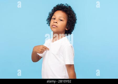 C'est moi. Portrait du garçon de curly préscolaire en T-shirt étant fier et arrogant se pointant vers lui avec une expression super-cilieuse sérieuse, chi égoïste Banque D'Images