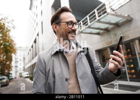 Image d'un homme d'affaires mûr et heureux dans des lunettes en utilisant le téléphone portable et en souriant tout en se tenant près de l'immeuble de bureaux dans la rue de la ville Banque D'Images