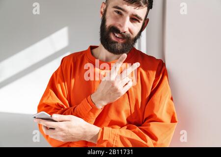 Image d'un jeune homme barbu portant une chemise orange montrant un signe de paix et tenant son smartphone tout en se tenant sur un mur blanc à l'intérieur Banque D'Images