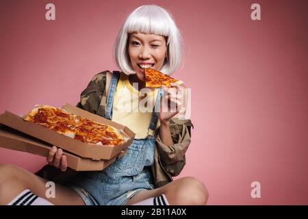 Image de la jeune fille asiatique portant une perruque blanche ayant amusant et manger de la pizza isolée sur fond rose Banque D'Images