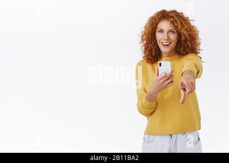 Fille satisfaite de votre pose comme photographier sur smartphone. Une jolie femme émouvante à tête rouge tient son téléphone mobile, pointant l'appareil photo avec un index Banque D'Images