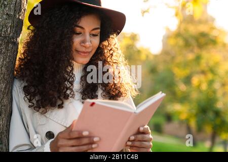 Belle femme africaine souriante portant un manteau d'automne penchant sur un arbre tout en se tenant au parc Banque D'Images