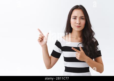 Une femme asiatique sceptique et sérieuse dans un t-shirt rayé semble suspecte et incertaine quant à l'offre douteuse, pointant vers le coin supérieur gauche, frognant et Banque D'Images