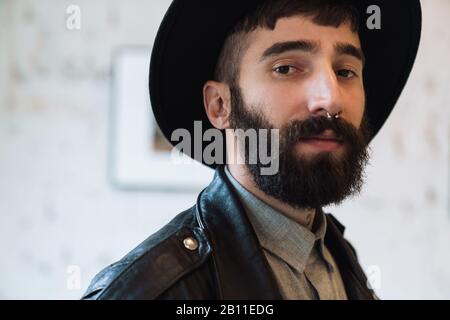 Photo closeup de jeune homme barbu portant chapeau posant et regardant l'appareil photo dans la salle lumineuse à l'intérieur Banque D'Images