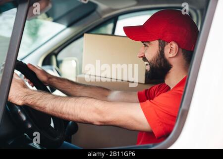 Image de l'homme de livraison barbu heureux en uniforme rouge souriant et camionnette avec des boîtes de colis Banque D'Images