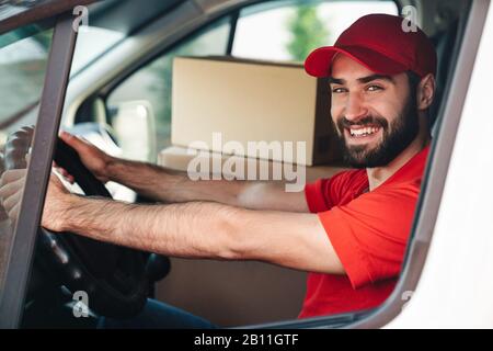 Image de l'homme de livraison barbu heureux en uniforme rouge souriant et camionnette avec des boîtes de colis Banque D'Images