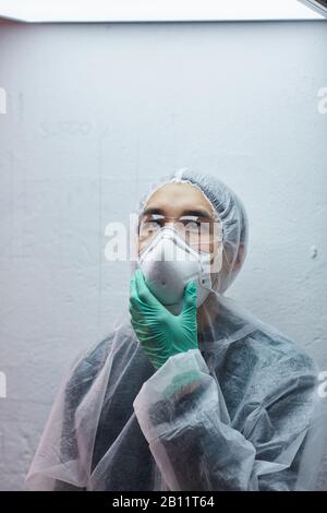 Jeune scientifique asiatique dans le masque et les lunettes de protection portant des gants de protection debout et regardant dans le laboratoire Banque D'Images