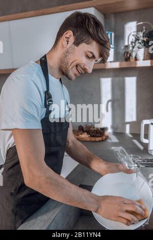 Smiling young man sort de la vaisselle propre lave-vaisselle Banque D'Images