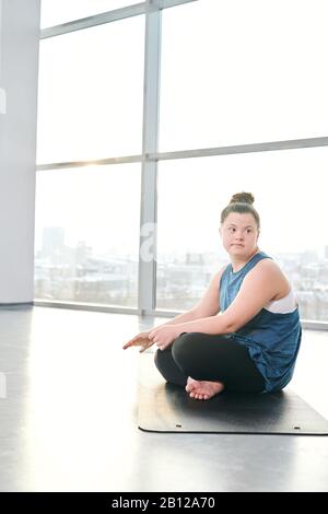 Jeune désactiver femme dans activewear traversant ses jambes tout en s'asseoir sur tapis dans la salle de gym Banque D'Images