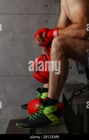 Gros plan sur un boxeur homme mi-adulte fatigué assis après un match d'entraînement, tenant des gants de boxe rouge avec les mains dans des enveloppements protecteurs de poignet, à faible angle Banque D'Images