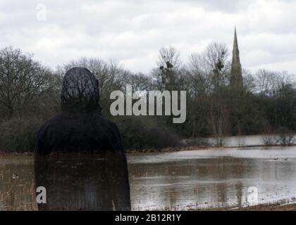 Une figure à capuche transparente, se tenant à côté d'un champ inondé, regardant une église voler. Une journée d'hivers de moody. Banque D'Images