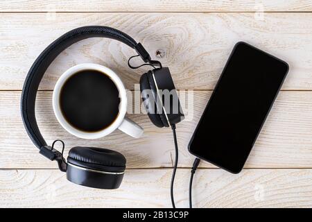 Smartphone, casque noir et tasse de café chaud sur une table en bois blanc. Appareil personnel moderne avec écran tactile pour la musique et la communication. Banque D'Images