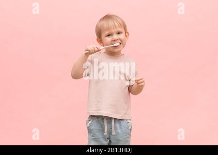 Enfant garçon blond brosse ses dents avec une brosse à dents sur un fond rose. Concept pour les articles sur les gencives saines, l'hygiène buccale et le traitement dans la dentistr Banque D'Images