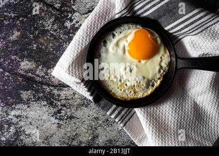 Côté ensoleillé, oeuf frit dans une poêle en fonte avec une serviette de cuisine rustique sur fond de béton. Vue de dessus Banque D'Images