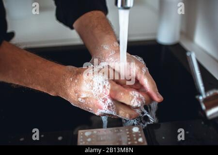 Couper la vue des mains fortes de l'homme les laver de la mousse de savon sous l'eau qui se poupe dans l'évier du robinet. Se tenir dans la cuisine avec les doigts de nettoyage. Mains voiture Banque D'Images