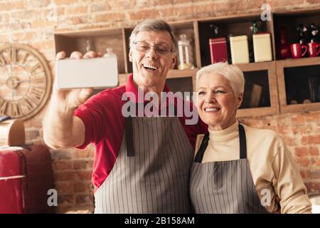 Couple âgé prenant selfie sur smartphone tout en cuisinant ensemble dans la cuisine Banque D'Images