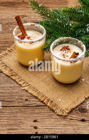 Cocktail d'eggnog dans deux verres disposés avec décoration de noël sur une vieille table en bois. Branche de sapin vert, neige artificielle, serviette en toile de sac, c Banque D'Images