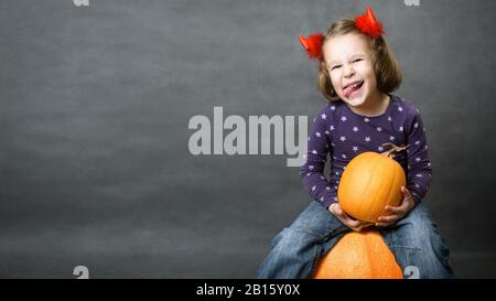 Fond d'Halloween avec adorable enfant et espace pour votre texte. Petite fille avec cornes de costume s'amuser. Un tout-petit mignon est assis sur la citrouille et montre à Banque D'Images