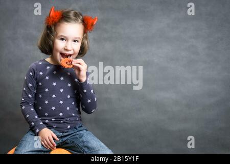 Fond d'Halloween avec adorable enfant et espace pour votre texte. Petite fille avec cornes de costume s'amuser. Les tout-petits sourient et mangent le tr d'Halloween Banque D'Images