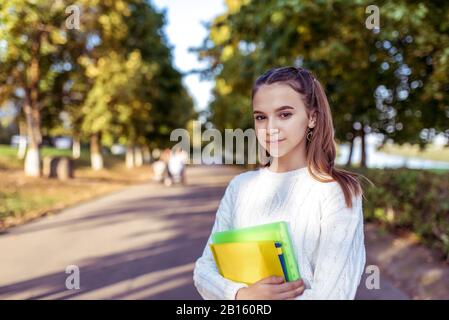 Fille écolière dans le parc d'été, dans les mains carnets manuels et livres. Arrière-plan route arbres vert herbe. Espace libre pour le concept de texte étudiant l'école Banque D'Images