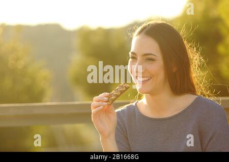 Une heureuse femme mangeant un bar à céréales assis sur un banc au coucher du soleil dans un parc Banque D'Images