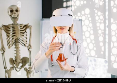 Portrait d'une jolie femme scientifique caucasienne dans un ballon de laboratoire contenant du liquide rouge, un échantillon sanguin ou une nouvelle substance, tout en portant des lunettes vr en laboratoire Banque D'Images