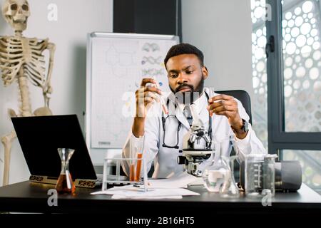 Beau jeune scientifique mâle africain virologue étudiant la réaction d'une substance chimique, d'un nouveau vaccin ou d'un médicament, tenant dans les mains deux tes Banque D'Images