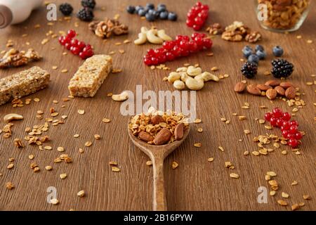 Spatule en bois avec granola à côté des baies, des noix, des flocons d'avoine et des barres de céréales sur fond en bois Banque D'Images