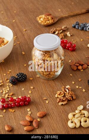 Pot de granola, bol et spatule à côté des amandes, noix de Grenoble, noix de cajou, baies sur fond de bois Banque D'Images