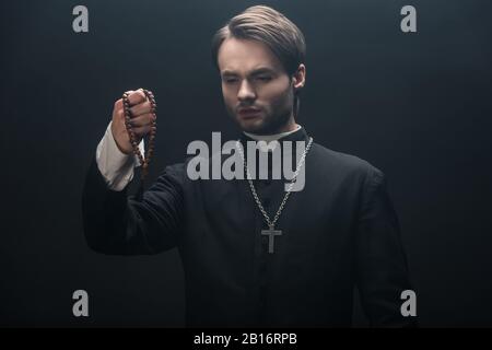 jeune prêtre catholique tendu regardant des perles rosaire en bois isolées sur le noir Banque D'Images