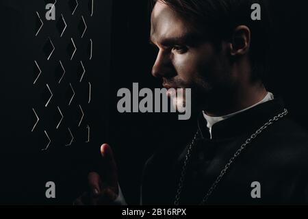 jeune prêtre catholique tendu regardant à travers la grille confessionnelle dans l'obscurité avec des rayons de lumière Banque D'Images