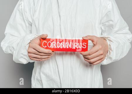 vue rognée de l'épidémiologiste dans un costume de hazmat portant une carte d'avertissement avec inscription au coronavirus sur fond gris Banque D'Images